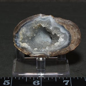 May include: A brown and gray geode with a hollow center filled with clear, sparkling quartz crystals, mounted on a clear acrylic stand positioned behind a ruler showing measurements in inches.