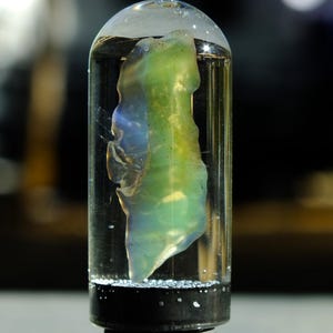 May include: A clear glass vial containing a translucent, abstract sculpture with green, blue, and yellow hues. The sculpture appears to be suspended in a clear liquid. The vial has a black base and a rounded top.