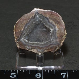 May include: A geode specimen with a triangular center of small, dark gray crystals surrounded by concentric bands of brown, tan, and white mineral layers. The geode is mounted on a clear acrylic stand positioned above a ruler showing inches.