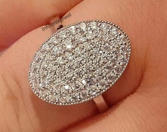 May include: A silver ring with an oval-shaped face covered in small, sparkling stones. The ring's band is thin, and the face is bordered by a delicate beaded design. The ring is shown on a person's finger.