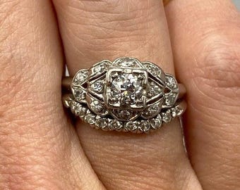 Könnte beinhalten: Ein Vintage-Diamant-Verlobungsring-Set mit einem Mittelstück, das von kleineren Diamanten umgeben ist. Der Ring ist in einer weißen Metallfassung gefasst.