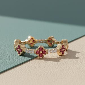 May include: A gold ring featuring a repeating clover design with red gemstones and small white gemstones. The ring is set against a neutral background, with the text "informixjewels" visible.