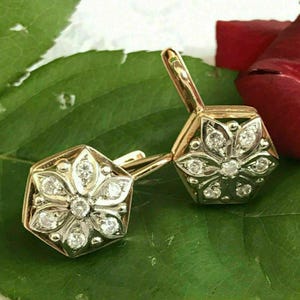 May include: A pair of gold earrings with a flower design set with clear gemstones. The earrings are on a green leaf background.