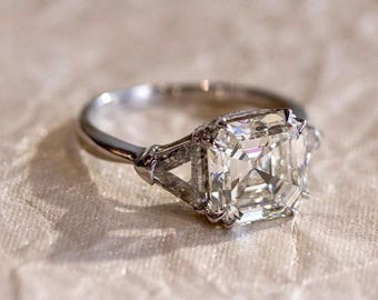 May include: A silver engagement ring with a large, square-cut diamond and two smaller triangular diamonds on either side.