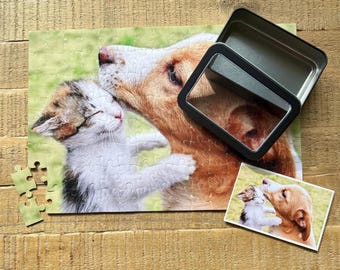Pode incluir: Um quebra-cabeça completo com um cão e um gatinho se abraçando, com uma imagem correspondente e uma caixa de metal. O quebra-cabeça mostra um cão marrom e branco com um gatinho calico. Várias peças do quebra-cabeça estão separadas.