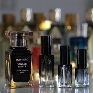 May include: A square glass perfume bottle labelled Tom Ford Vanille Fatale Eau de Parfum 50 ml, positioned next to three smaller, clear glass spray vials with black caps. The main bottle features a dark liquid and a metallic silver-toned cap.