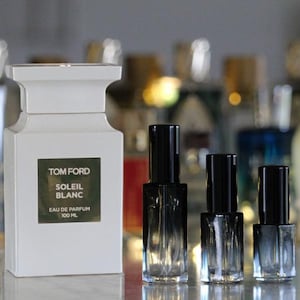 May include: A white rectangular Tom Ford Soleil Blanc Eau de Parfum 100 ml bottle with a dark label, positioned next to three smaller, empty, clear glass spray bottles with black caps, arranged in a row from tallest to shortest.