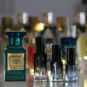 May include: A rectangular teal glass perfume bottle labeled Tom Ford Azure Lime Eau de Parfum 50 ml, positioned next to three smaller, clear glass spray vials with black caps, all arranged in a row against a blurred background of other bottles.