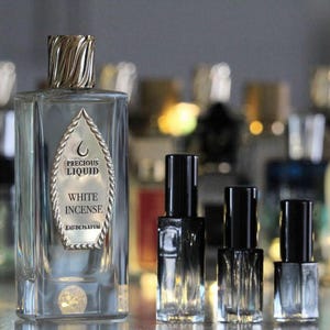 Peut inclure: Un flacon de parfum en verre transparent avec un bouchon torsadé doré et une étiquette en forme de bouclier portant l'inscription Precious Liquid White Incense Eau de Parfum, accompagné de trois flacons plus petits à bouchons noirs.