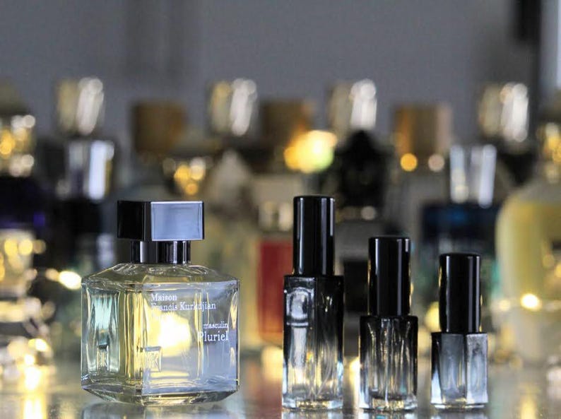 Op de afbeelding: Vijf glazen flessen parfum met zwarte dopjes. De fles in het midden is groter en heeft de tekst "Maison Francis Kurkdjian masculin Pluriel" op het etiket.