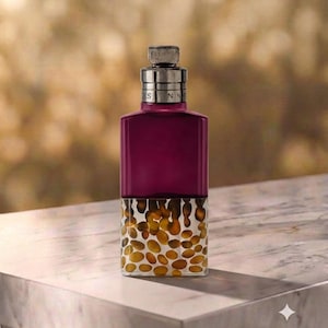 May include: A rectangular glass perfume bottle with a deep magenta upper section and a clear lower section filled with amber-coloured, pebble-like shapes. The bottle features a metallic silver cap with the text "DRIES VAN NOTEN" engraved on it.