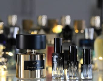 May include: A large gold perfume bottle with a black cap and a black label reading Kirke Tiziana Terenzi, positioned next to three smaller, clear glass perfume sample vials with black caps, all featuring a cylindrical design and metallic accents.