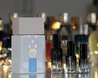 May include: A large white rectangular perfume bottle with a light wood cap and a blue label reading Bianco Latte 100 ml 3.4 fl oz, accompanied by three smaller clear glass spray bottles with black caps arranged in a row to the right side.
