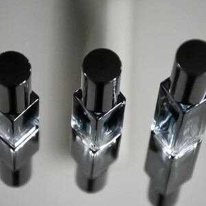 Op de afbeelding: Drie kleine, vierkante glazen flesjes met zwarte dopjes. De flesjes zijn gevuld met een heldere vloeistof en zijn in een rij op een reflecterend oppervlak gerangschikt.