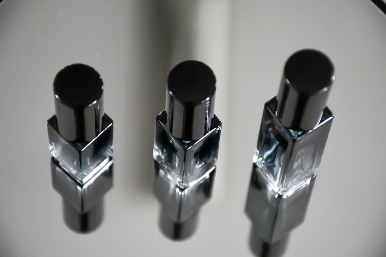 Op de afbeelding: Drie kleine, vierkante glazen flesjes met zwarte dopjes. De flesjes zijn gevuld met een heldere vloeistof en zijn in een rij op een reflecterend oppervlak gerangschikt.