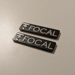 Può includere: Due badge rettangolari neri Focal con testo e logo bianchi. La parola "FOCAL" è in un font sans-serif. Il logo è un design stilizzato sopra la parola. I badge sono su una superficie chiara.