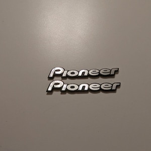 Puede incluir: Dos emblemas Pioneer plateados con contorno negro sobre un fondo gris claro. La palabra "Pioneer" está escrita en una fuente estilizada. Los emblemas son probablemente para audio de coche u otros dispositivos electrónicos.