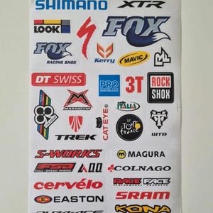 May include: A sheet of bicycle component brand stickers, including Shimano, Fox, DT Swiss, Trek, and SRAM. The stickers feature various logos in red, blue, white, and yellow. The sheet is on a white background.