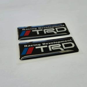 May include: Two rectangular black adhesive emblems featuring the silver text Racing Development and the acronym TRD in a large silver font. Each emblem includes a small graphic element with a blue stripe and a red stripe on the left side.