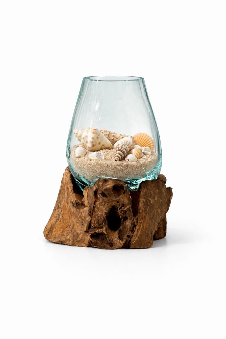 May include: A clear glass vase filled with sand and seashells, resting on a natural wood base. The vase has a teardrop shape and contains various seashells in different colors and sizes. The wood base is a rough, organic shape.