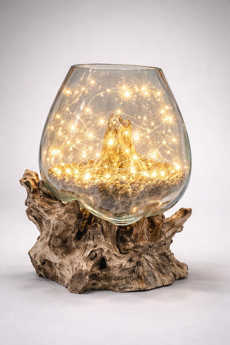 May include: A decorative glass terrarium with warm white fairy lights inside, resting on a natural wood base. The glass vessel is filled with small stones and a piece of driftwood. The overall design creates a warm, inviting ambiance.