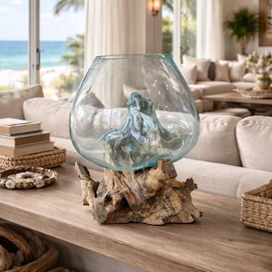 May include: A large, clear glass bowl with a wide opening, resting on a natural, weathered wood base. The bowl contains a blue-tinted, organic-shaped glass element. The decorative piece sits on a wooden table in a bright, airy living room.