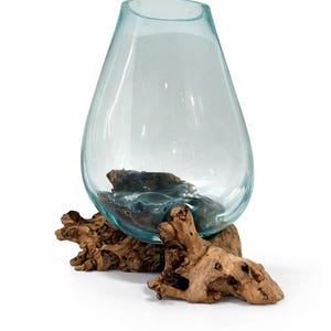 May include: A clear, teardrop-shaped glass vase with a light blue tint, resting on a natural, weathered driftwood base. The vase is empty, showcasing its smooth surface and the organic texture of the wood.