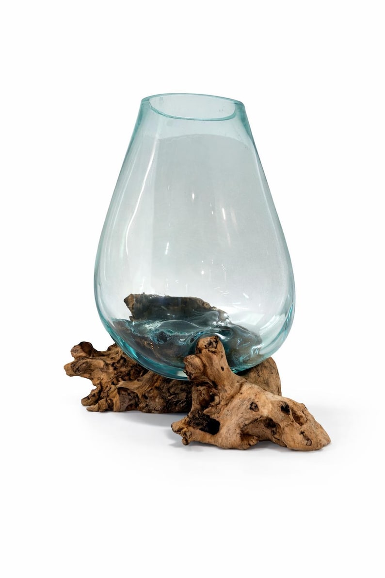 May include: A clear, teardrop-shaped glass vase with a light blue tint, resting on a natural, weathered driftwood base. The vase is empty, showcasing its smooth surface and the organic texture of the wood.