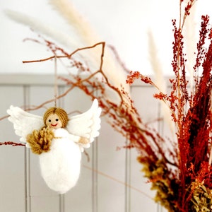 Puede incluir: Un adorno de &aacute;ngel de fieltro blanco con cabello rubio y una flor de fieltro marr&oacute;n en su mano. El &aacute;ngel est&aacute; colgado de una rama con flores secas en el fondo.