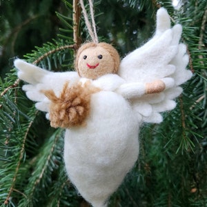 Puede incluir: Un adorno de &aacute;ngel de fieltro blanco con alas de fieltro marr&oacute;n y una cara sonriente. El &aacute;ngel sostiene una bola de fieltro marr&oacute;n en sus manos.