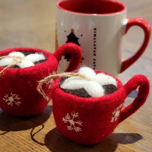 Puede incluir: Dos adornos de taza de fieltro rojo con bordado de copo de nieve blanco y cobertura de malvavisco blanco. Detrás de los adornos hay una taza de cerámica con interior y asa rojos. La taza tiene un diseño de árbol de Navidad.