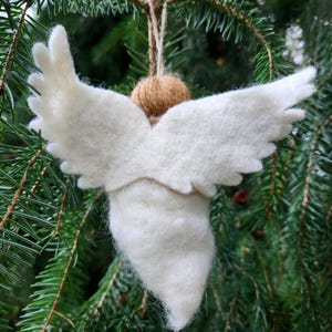 Puede incluir: Un adorno de &aacute;ngel de fieltro blanco con alas, colgado de una cuerda de yute marr&oacute;n. El adorno est&aacute; colgado de una rama de pino verde.