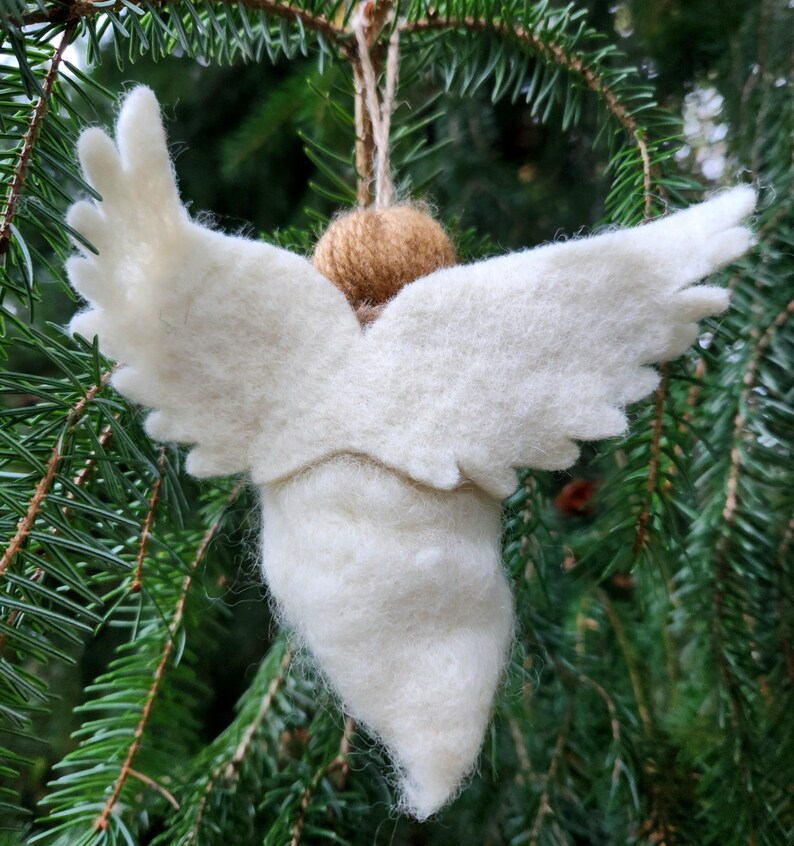 Puede incluir: Un adorno de &aacute;ngel de fieltro blanco con alas, colgado de una cuerda de yute marr&oacute;n. El adorno est&aacute; colgado de una rama de pino verde.