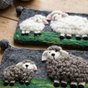 May include: Two rectangular felt pouches in dark gray, each featuring a needle-felted scene of two sheep standing on a green grassy patch. The top pouch displays white sheep, while the bottom pouch features brown sheep, both with pom-pom zipper pulls.