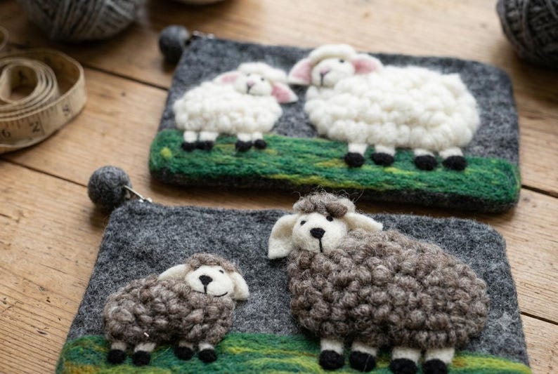 May include: Two rectangular felt pouches in dark gray, each featuring a needle-felted scene of two sheep standing on a green grassy patch. The top pouch displays white sheep, while the bottom pouch features brown sheep, both with pom-pom zipper pulls.