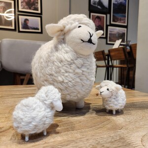 May include: Three white sheep figurines of varying sizes are arranged on a wooden surface. The sheep have fluffy, textured wool and black facial details. The largest sheep stands upright, while the smaller ones are positioned nearby.