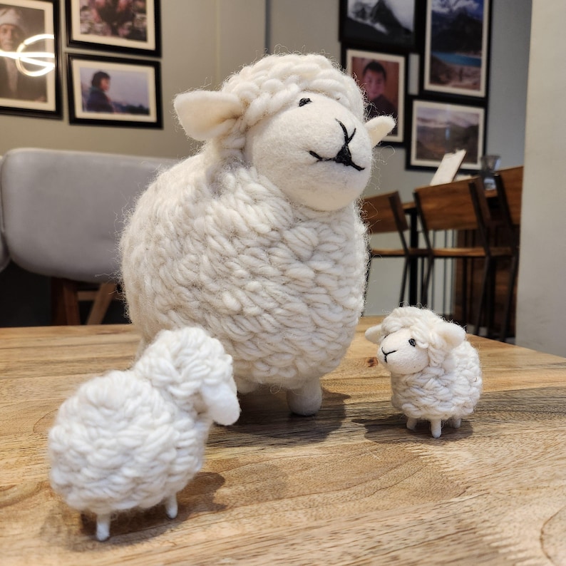May include: Three white sheep figurines of varying sizes are arranged on a wooden surface. The sheep have fluffy, textured wool and black facial details. The largest sheep stands upright, while the smaller ones are positioned nearby.