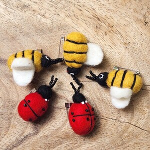 May include: A collection of handmade felt insect pins, including bees and ladybugs. The bees are yellow and black with white wings, while the ladybugs are red with black spots. Each pin has a safety pin backing.