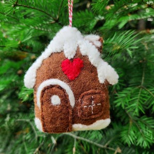 May include: A brown felt gingerbread house Christmas ornament with white accents and a red heart. The ornament is hanging from a red and white string against a backdrop of green pine needles.