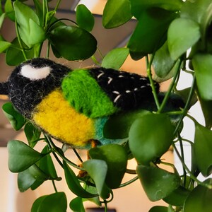 May include: A hand-felted bird made from black, white, yellow and green wool. The bird is perched on a green leafy plant.