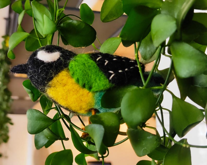 May include: A hand-felted bird made from black, white, yellow and green wool. The bird is perched on a green leafy plant.