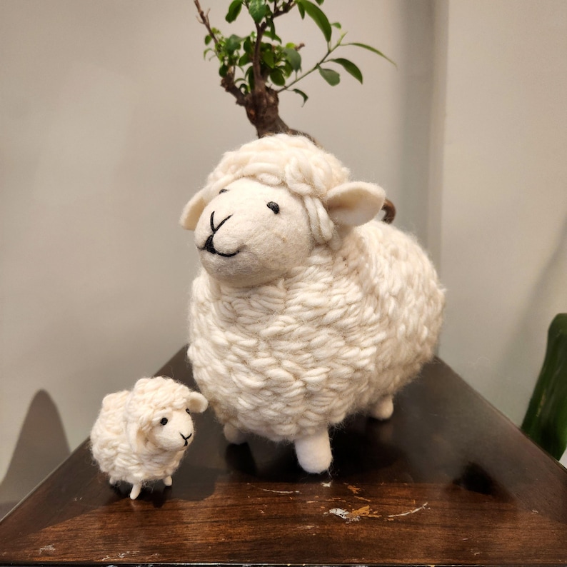 May include: Two white wool sheep figurines, one large and one small, are displayed on a dark wooden surface. The sheep have black eyes and a stitched smile. A small bonsai tree is in the background.