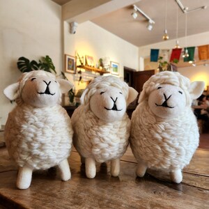 May include: Three handcrafted white sheep figurines with black facial details and small ears. The sheep are made of a textured, wool-like material and stand on four small legs. They are arranged on a wooden surface.