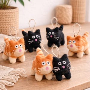 Peut inclure: Ornements de chats faits à la main en feutre. Les chats sont de couleur noire et orange tigré, avec des moustaches blanches et des nez roses. Ils sont suspendus par une ficelle en jute. Les chats sont posés sur une surface en bois.