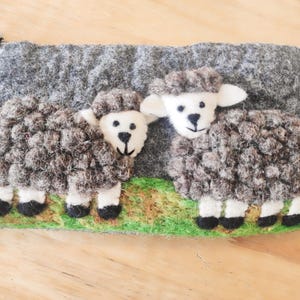 May include: A rectangular felt pouch featuring two needle-felted sheep with textured dark gray wool bodies, white faces, and black hooves. The sheep stand on a green grassy base against a dark gray background, with a small white zipper pull attached.