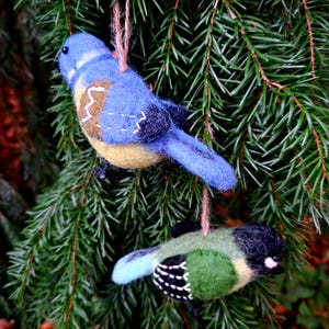 May include: Two felt bird decorations hanging from a pine tree branch. The larger bird is blue with yellow accents and the smaller bird is green with black accents.