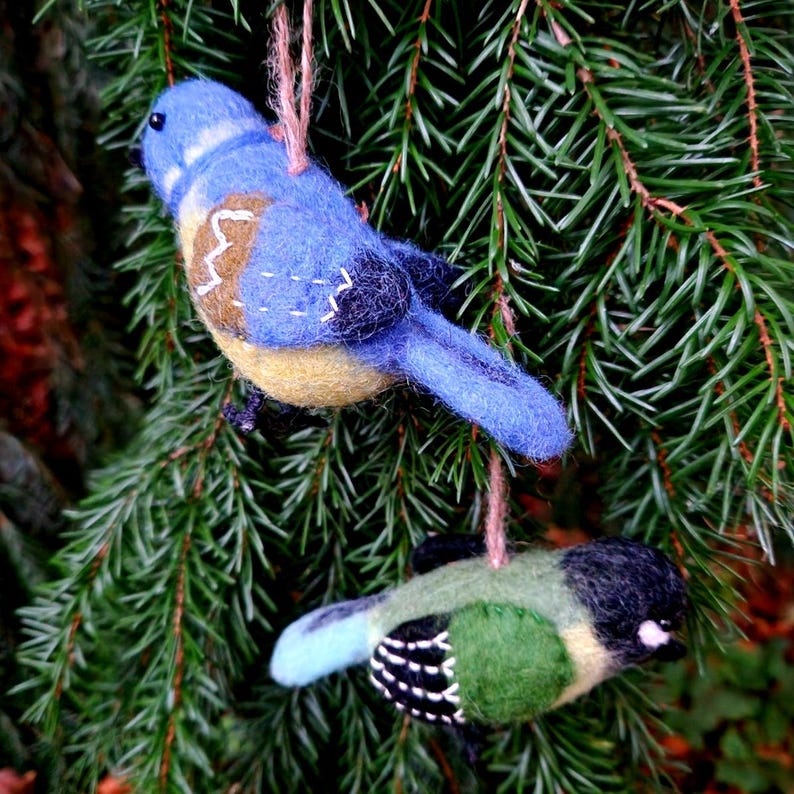 May include: Two felt bird decorations hanging from a pine tree branch. The larger bird is blue with yellow accents and the smaller bird is green with black accents.