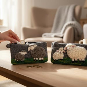 May include: Two rectangular felt pouches featuring textured sheep appliqu&eacute;s on a green field. One pouch is dark gray with gray sheep, and the other is dark gray with white sheep. Both have a zipper closure and a small round pull tab on the side.