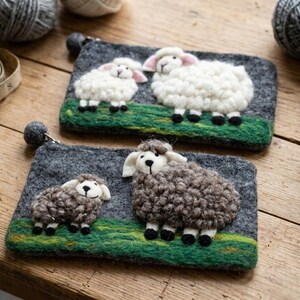 May include: Two rectangular felt pouches featuring needle-felted sheep designs. The top pouch displays two white sheep on a green field against a dark gray background. The bottom pouch features two brown sheep on a green field against a dark gray background.