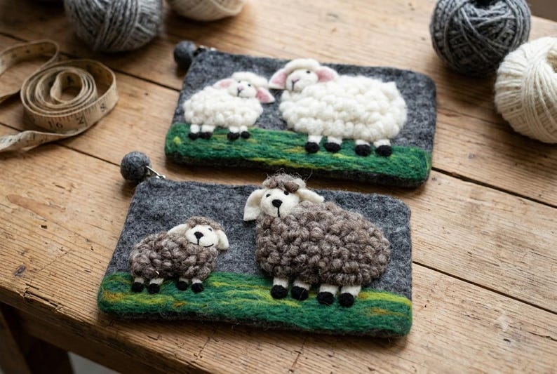 May include: Two rectangular felt pouches featuring needle-felted sheep designs. The top pouch displays two white sheep on a green field against a dark gray background. The bottom pouch features two brown sheep on a green field against a dark gray background.